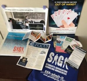 Promotional display of gambling awareness materials: brochures, flyers, cards, and an 'Out of Hand' card game on a table, with a blue SAGA bag nearby.