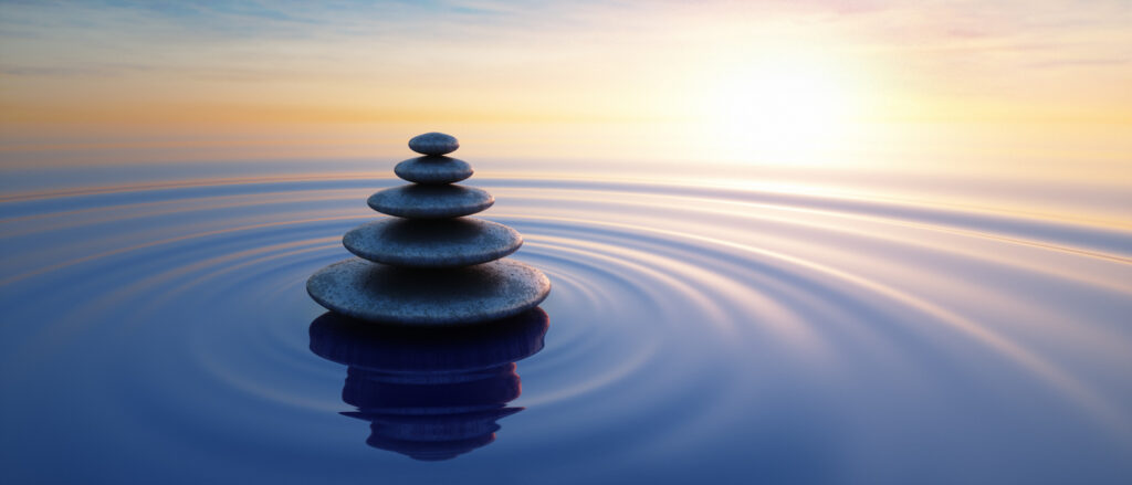 Stack of six smooth stones balanced in a tall tower on calm water, with circular ripples and a warm sunset glow on the horizon reflecting in the surface.